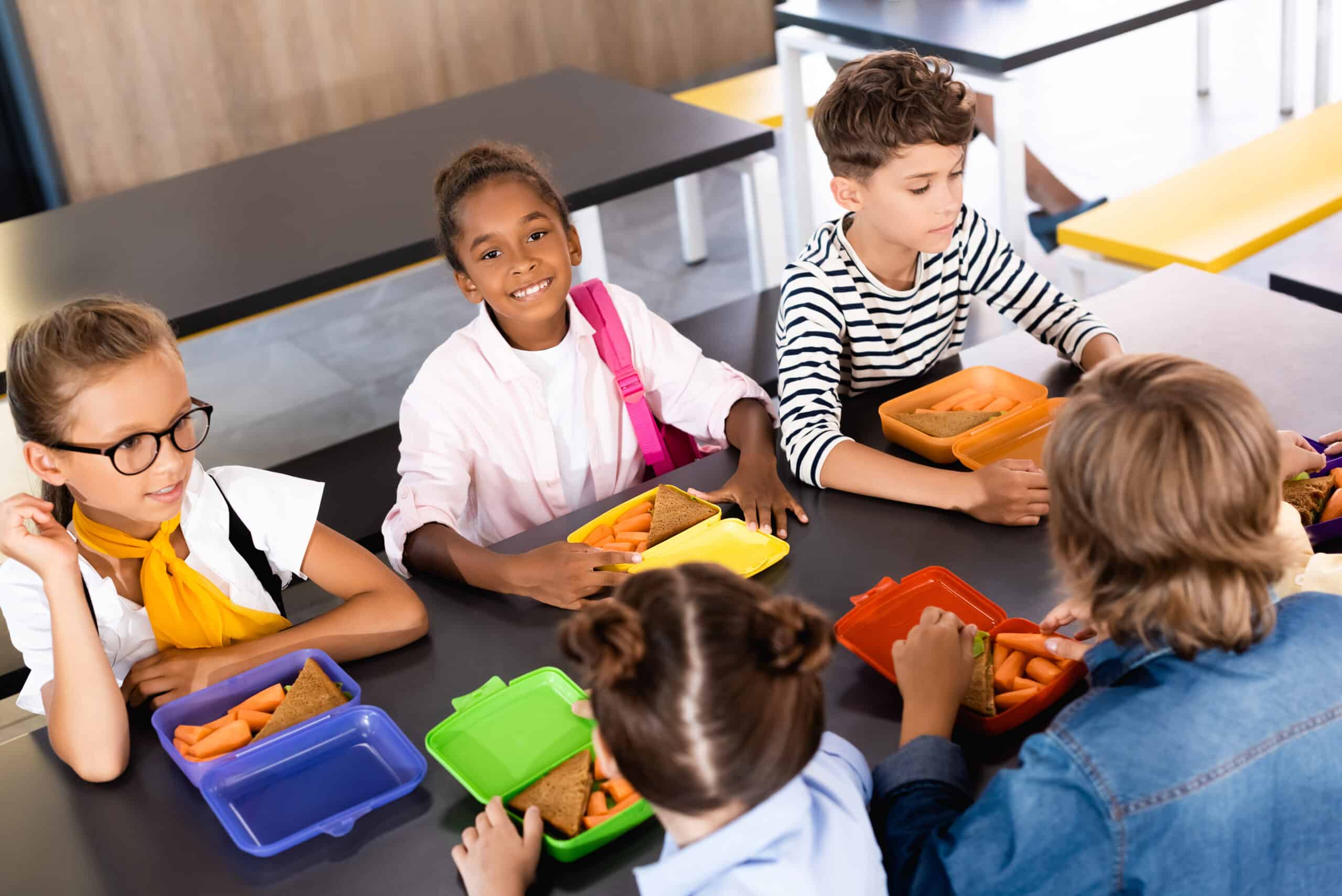 6 kinderen die in een schoolkantine brood en worteltjes eten uit hun eigen felgekleurde trommeltje. Deze leerlingen zitten in de schoolkantine.